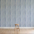 A simple wooden stool sits on a light wooden floor in front of a wall covered with Painted Stripe Wallpaper, Blue, featuring hand-painted vertical blue and white irregular stripes.