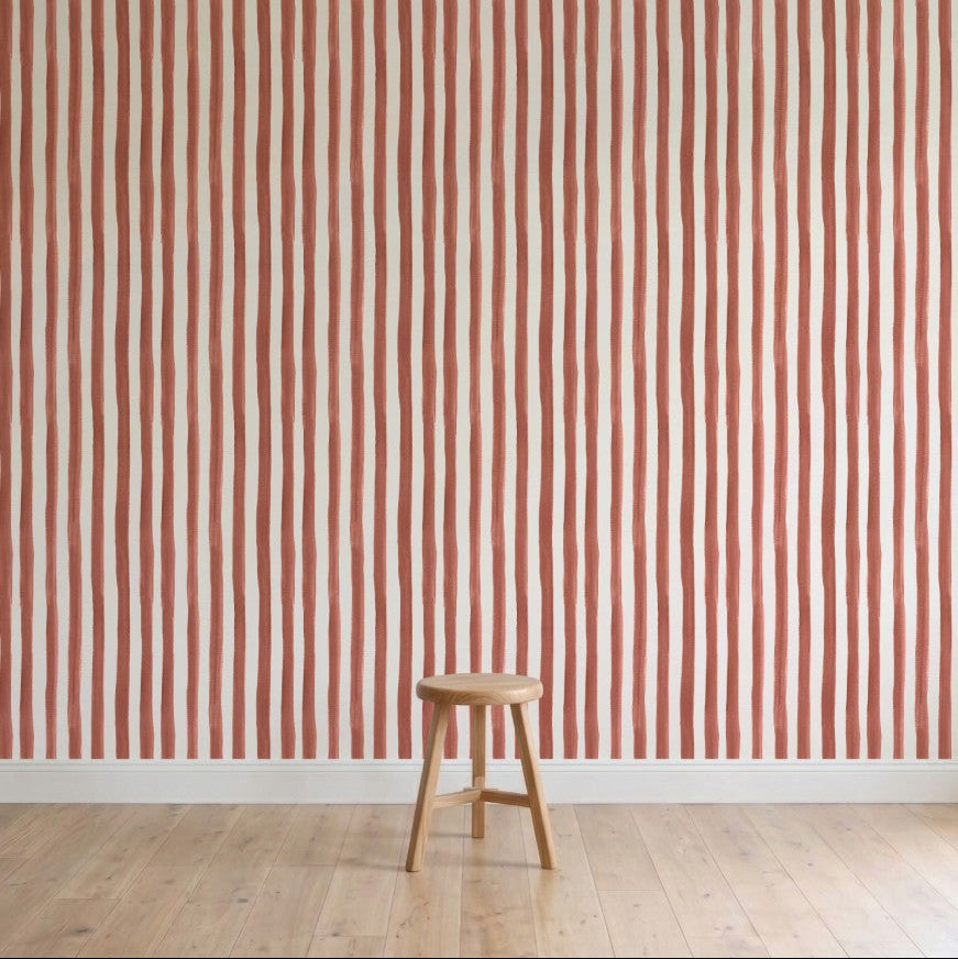 A simple wooden stool is on a light wood floor in front of a wall decorated with Painted Stripe Wallpaper, Red, featuring hand-painted wavy red and white patterns.