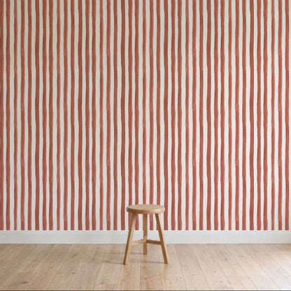 A simple wooden stool is on a light wood floor in front of a wall decorated with Painted Stripe Wallpaper, Red, featuring hand-painted wavy red and white patterns.