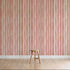 A simple wooden stool is on a light wood floor in front of a wall decorated with Painted Stripe Wallpaper, Red, featuring hand-painted wavy red and white patterns.
