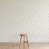 A plain wooden stool sits on a light wood floor before a cream-colored wall decorated with Soaring Hearts Wallpaper featuring subtle, muted bird designs. The minimalist scene appears calm and uncluttered.