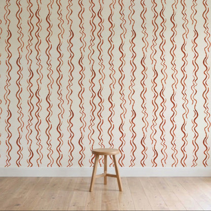 A wooden stool sits on a light wood floor in front of a cream wall featuring Wiggle Stripe Wallpaper, Red—vertical wavy red and beige stripes. The minimal, well-lit setting highlights this stylish children’s wallpaper.