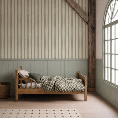 A cozy wooden bed with a checkered quilt sits in a light-filled room featuring Scallop Stripe Wallpaper in Green, wainscoting, a large arched window, and a woven basket on the floor.