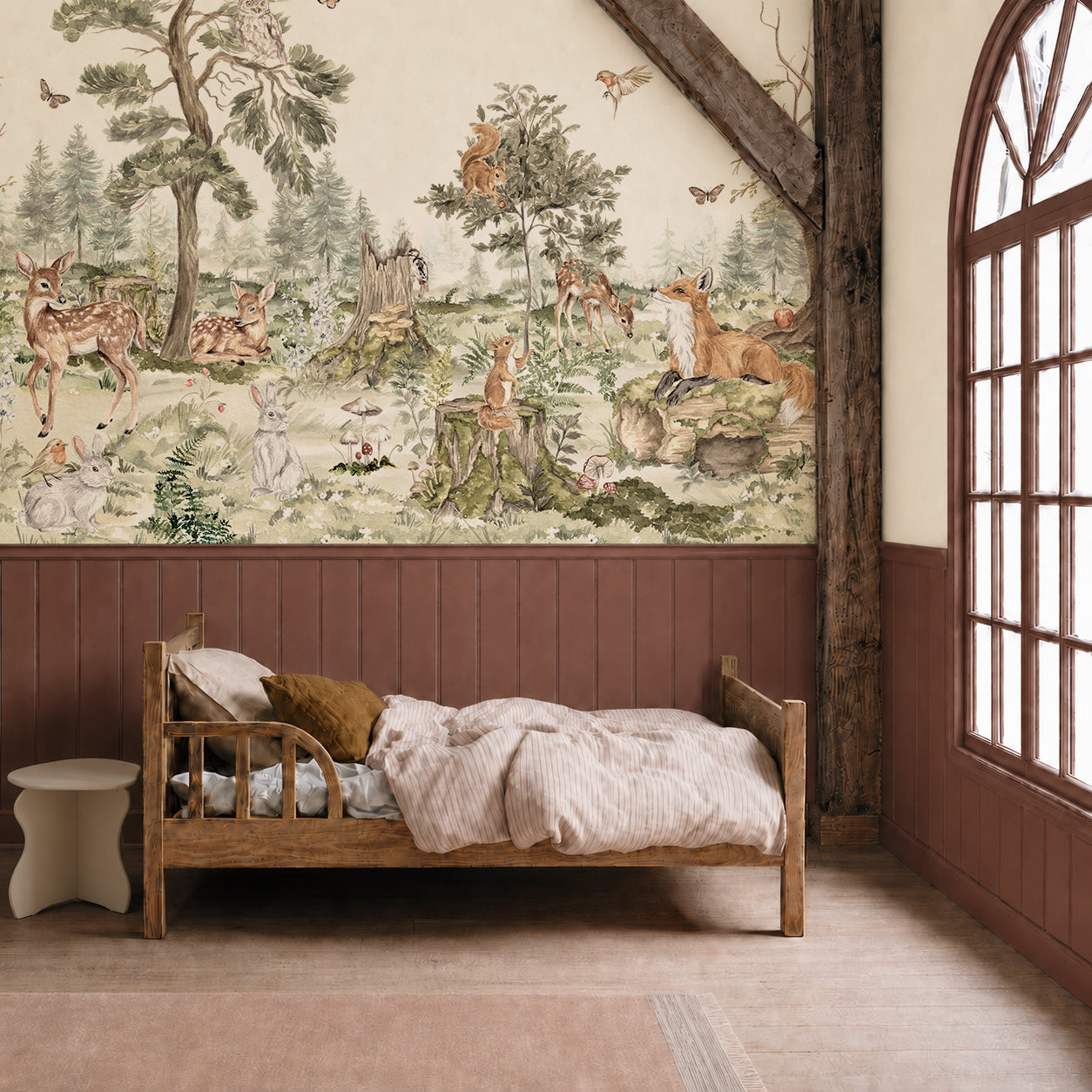 A cozy children’s bedroom features a wooden bed, striped bedding, and the enchanting “Into The Forest Mural” on the wall, depicting deer, foxes, rabbits, and trees. Sunlight pours in through a large arched window, illuminating the hand-painted mural and bringing the magical woodland scene to life.