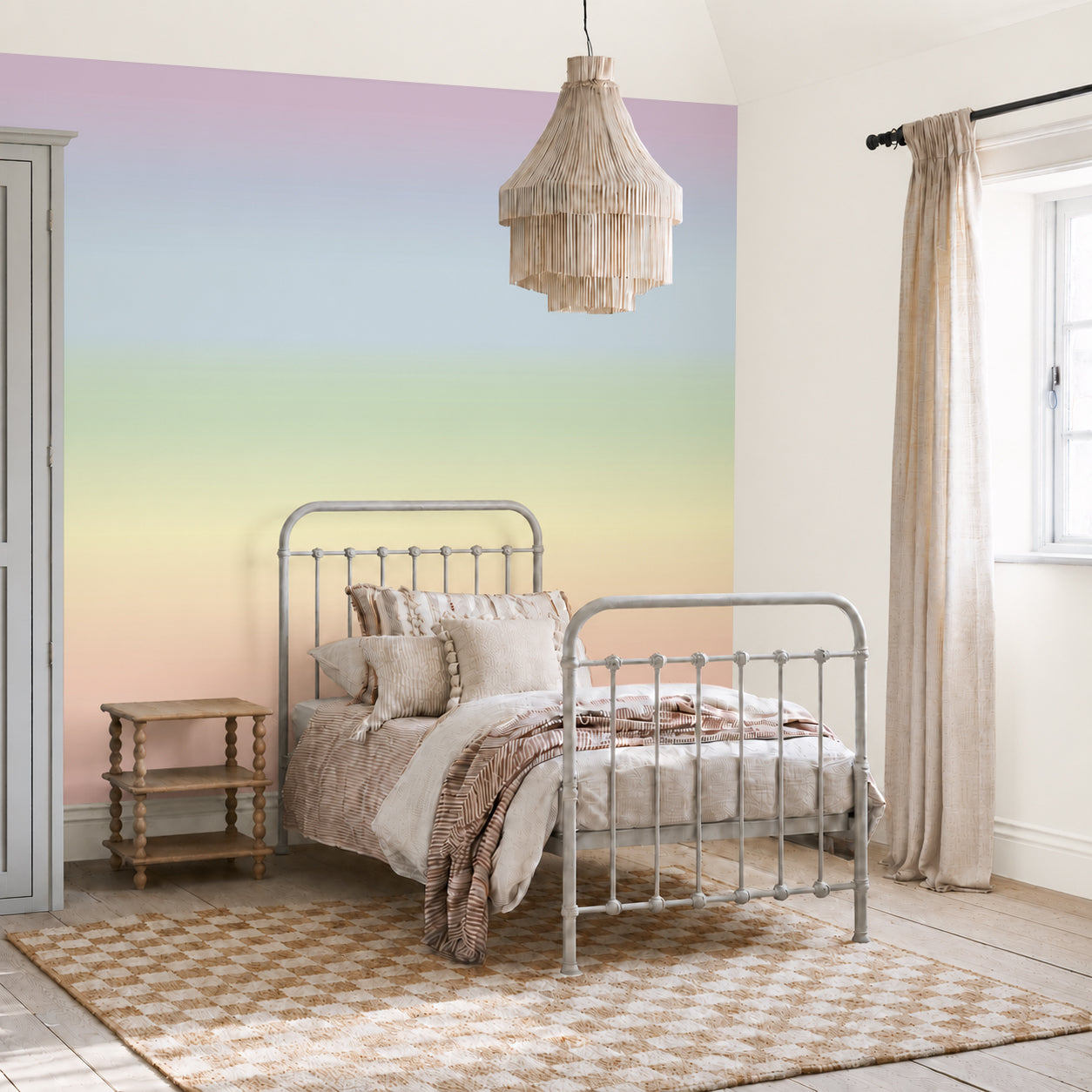 Cozy bedroom with a vintage metal bed, neutral bedding, a pastel gradient wall featuring the Ombre Mural, Rainbow wallpaper, a woven rug, a wooden side table, a fringed light fixture, and a window with beige curtains letting in natural light.