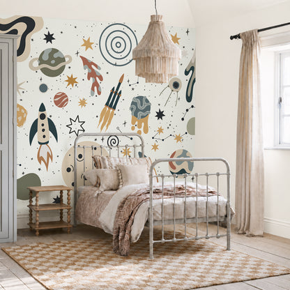 A cozy bedroom features a metal-framed bed with beige bedding, wooden side tables, and a window. The wall is adorned with the To The Moon Mural—a vibrant space-themed wallpaper showcasing rockets, planets, and stars—perfect for inspiring young imaginations. A woven pendant light hangs from the ceiling, making this mural an ideal kids room wallpaper choice.