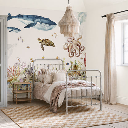 A cozy bedroom features a metal bed and woven rug, with light streaming through beige curtains to illuminate the Under The Sea Mural, Watercolour. This stunning wallpaper displays a captivating ocean scene complete with a whale, turtle, octopus, fish, and coral, bringing an enchanting under-the-sea feel to the room.