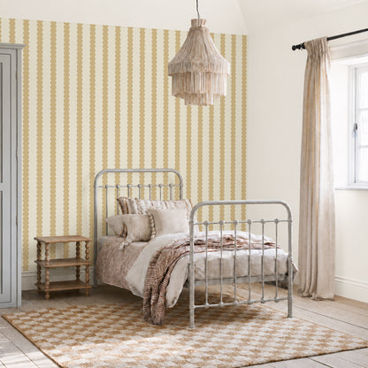 A cozy bedroom with a metal bed, neutral bedding, a wooden side table, a fringed pendant lamp, Scallop Stripe Wallpaper in Yellow on the walls, and a window with beige curtains letting in natural light. A patterned rug lies on the wooden floor.