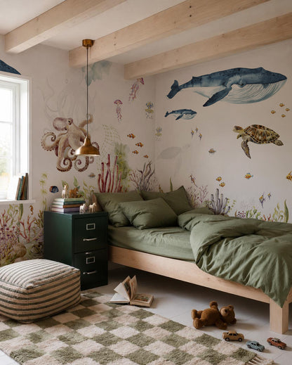 A cozy bedroom featuring green bedding, a checkered rug, books, and ocean-themed wall art with a whale, turtle, octopus, and fish—including an accent wall adorned with the Under The Sea Mural, Watercolour. Sunlight streams in through a window next to a green filing cabinet and a striped pouf.