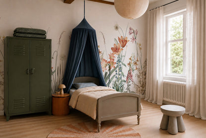 Children’s bedroom featuring a gray bed under a blue canopy, Quinns Flower Field Mural as wall art, green locker-style wardrobe, round side table with lamp, circular rug, large window with white curtains, and wooden stool.