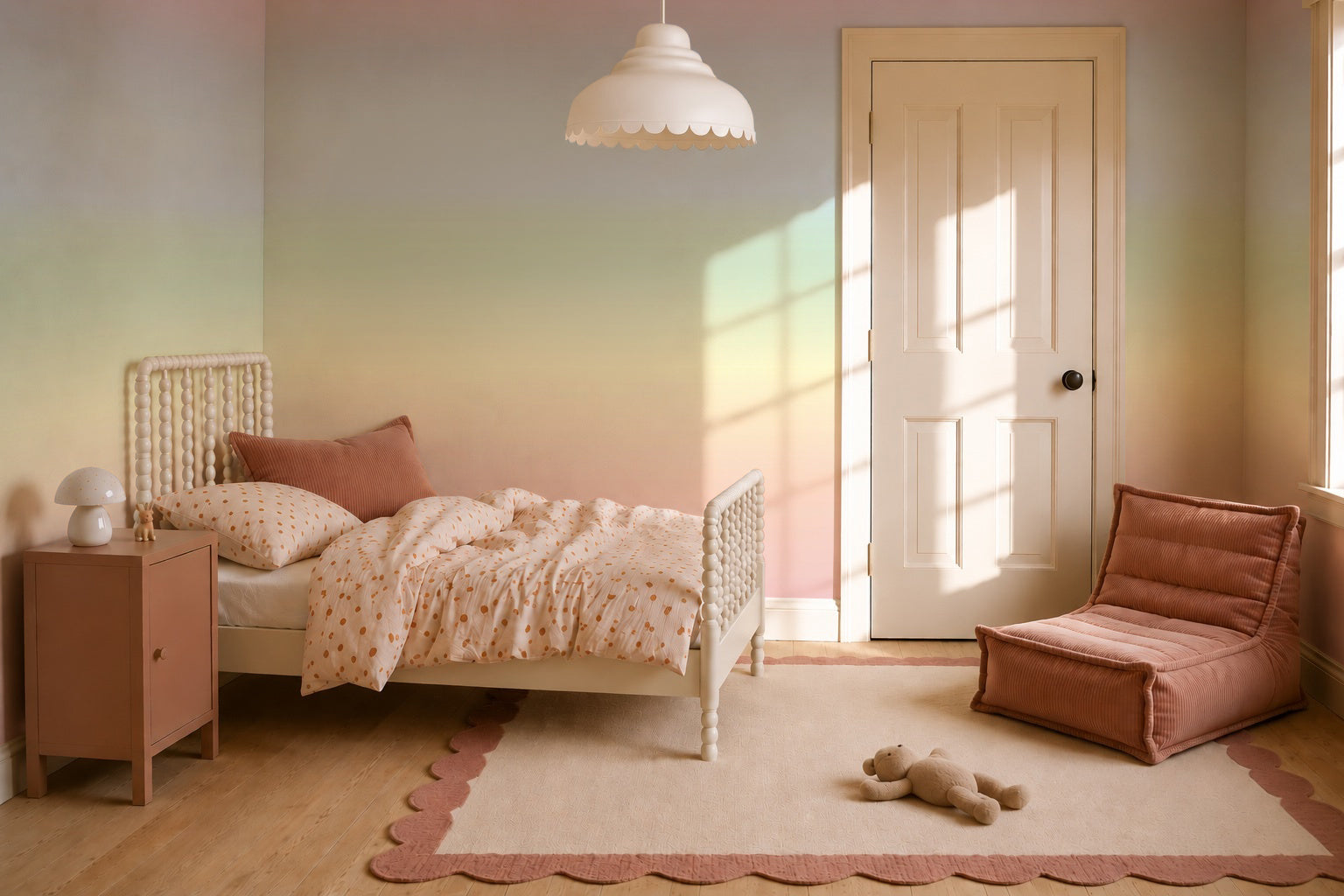 A cozy kids bedroom featuring the Ombre Mural, Rainbow as a pastel ombre wallpaper mural, a white bed, scalloped rug, rust-colored cushion seat, small nightstand with lamp, and a plush teddy bear on the floor. Sunlight streams in through a window.