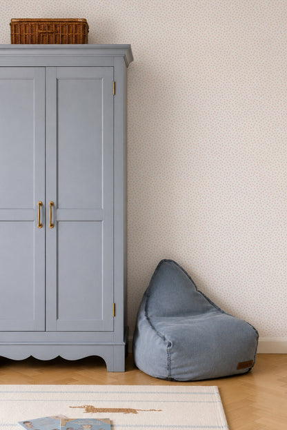 A light blue armoire with brass handles stands beside a blue bean bag chair. A brown wicker basket sits on top. The room features whimsical wall decor and a white rug with blue accents on a wooden floor, all set against Sprinkle Wallpaper in pink.