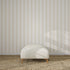 A cream-colored ottoman with wooden legs rests on a woven carpet in front of Scallop Stripe Wallpaper Cloud, as soft sunlight and window shadows play across the wall and floor.