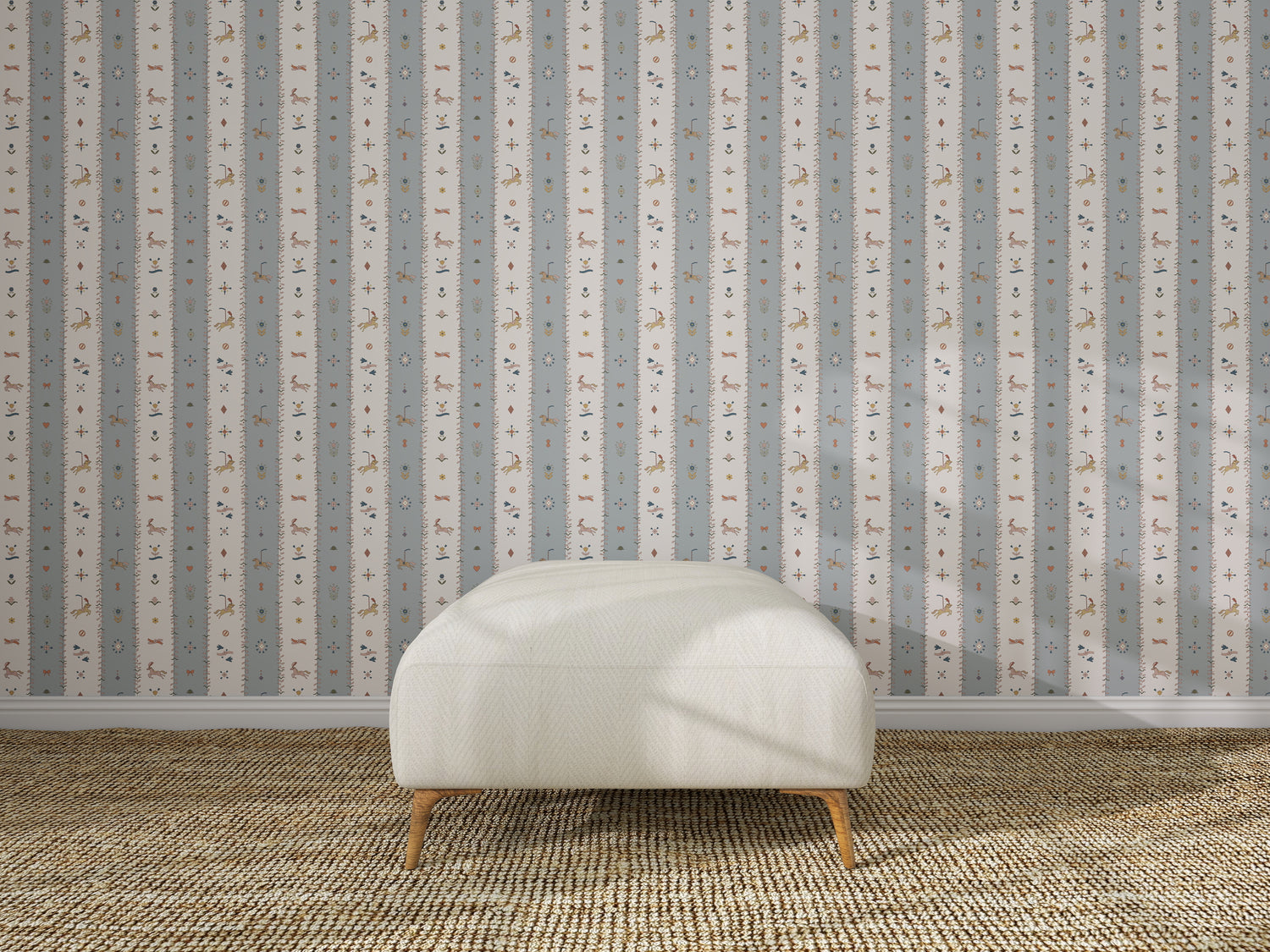 A white upholstered bench with wooden legs sits on a woven tan rug in front of The Little Carousel Stripe Wallpaper, Blue, featuring vertical stripes adorned with small animal and plant motifs for a playful touch.