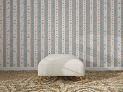 A white upholstered bench with wooden legs sits on a woven tan rug in front of The Little Carousel Stripe Wallpaper, Blue, featuring vertical stripes adorned with small animal and plant motifs for a playful touch.