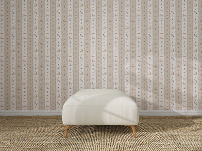 A white upholstered bench with wooden legs sits on a textured beige rug in front of The Little Carousel Stripe Wallpaper, Rose, featuring vertical stripes and delicate floral motifs.