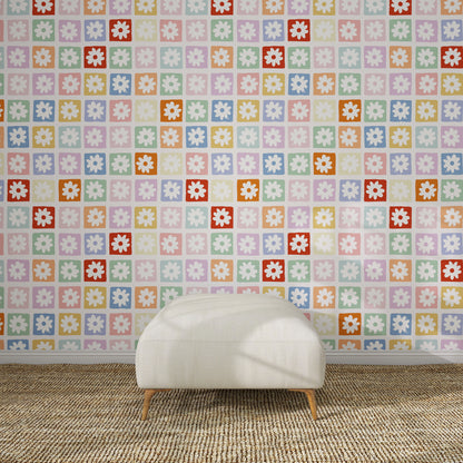 A white, cushioned ottoman sits on a textured brown rug in front of a wall decorated with the Floral Fun land Repeat Wallpaper Pattern—a vibrant retro floral design in pastel pinks, purples, blues, yellows, oranges, reds, and greens.