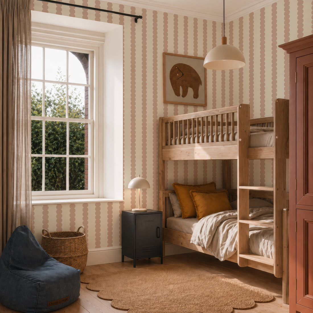 A cozy kids’ bedroom with wooden bunk beds, earth-toned bedding, a black nightstand, a round lamp, a blue beanbag, wicker basket, elephant wall art, window with curtains, and Scallop Stripe Wallpaper in Pink.