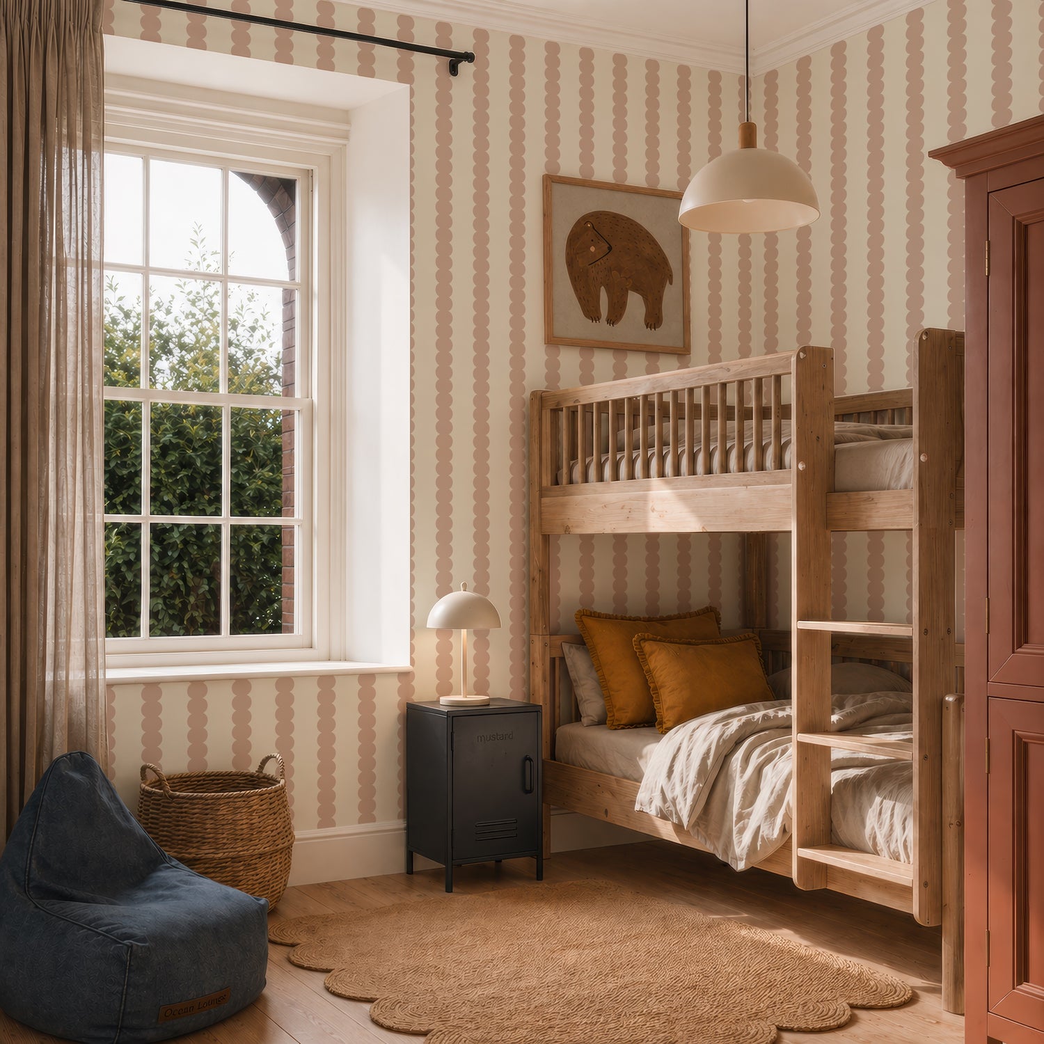A cozy kids’ bedroom with wooden bunk beds, earth-toned bedding, a black nightstand, a round lamp, a blue beanbag, wicker basket, elephant wall art, window with curtains, and Scallop Stripe Wallpaper in Pink.