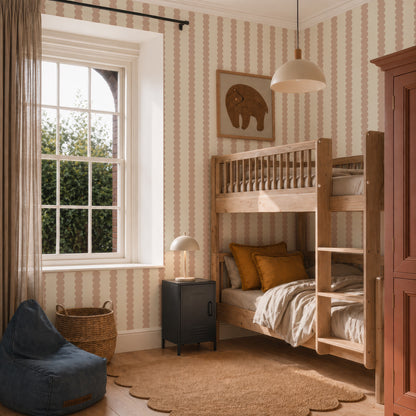 A cozy kids’ bedroom with wooden bunk beds, earth-toned bedding, a black nightstand, a round lamp, a blue beanbag, wicker basket, elephant wall art, window with curtains, and Scallop Stripe Wallpaper in Pink.