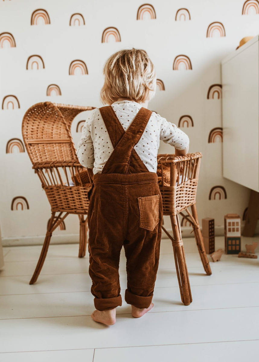 A blonde toddler in a white polka-dotted shirt and brown overalls stands barefoot in a room decorated with Raining Rainbows Wallpaper Repeat Pattern, facing two wicker chairs and a small table filled with wooden toys.