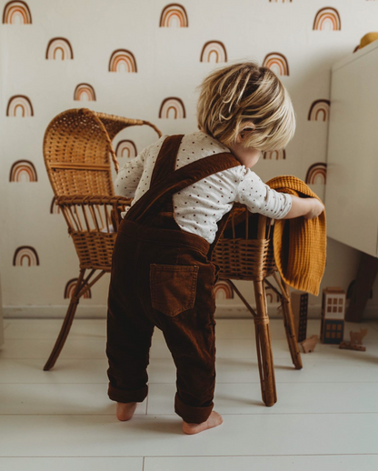 A blond toddler in brown overalls and a white polka dot shirt places an orange blanket on a wicker chair in a room with light floors and walls adorned with the whimsical Raining Rainbows Wallpaper Repeat Pattern.