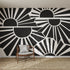 A modern room features the striking Sunbeams Wallpaper Repeat Pattern | Charcoal with geometric black and white stylized suns and rays, alongside a wooden chair with a cushion and a woven rug on the floor.