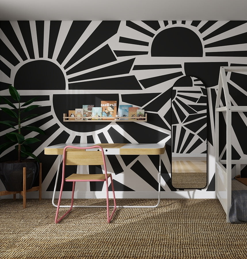 A modern workspace features a wooden desk, pink chair, books on a shelf, tall plant, and bold Sunbeams Wallpaper Repeat Pattern | Charcoal. A mirror reflects the wallpaper’s striking sunburst design.