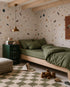 A cozy bedroom with Terrazzo Spritz Mural, a wooden bed with green bedding, a green nightstand topped with animal figurines, a striped pouf, a checkered rug, and toys scattered across the floor.