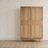 The Thank My Lucky Stars Wallpaper Sample adds a delicate touch, with its cream color and subtle green floral pattern, enhancing the quality and charm of the space behind a simple wooden wardrobe on a light wood floor.