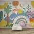 The Happy Wall Wallpaper Mural showcases colorful shapes, plants, stars, rainbows, a crescent moon, and playful patterns on a white wall behind a white ottoman with a woven rug, creating a lively and whimsical atmosphere.