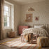 A cozy, sunlit bedroom features vibrant Powdered Petals Wallpaper, a white metal bed, pink dresser, woven basket, pouf, soft striped rug, framed dog art, and a letter A wall hanging for a warm and inviting feel.