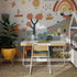 A small wooden desk with a white chair stands on a woven rug before the Wonderpark Wallpaper Mural—a colorful, whimsical rainbow scene with trees, boats, and playful shapes. A potted plant is placed to the left of the desk.