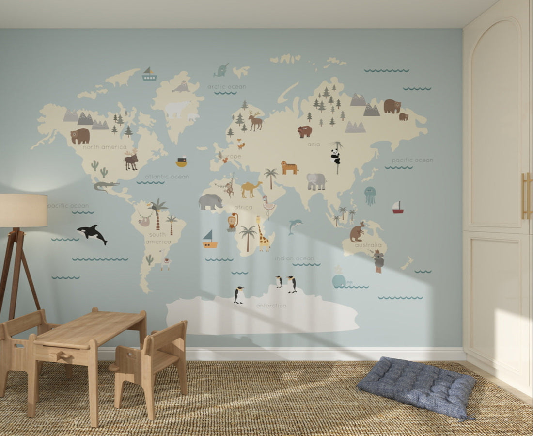 A childrens playroom for a young explorer, with the Character World Map Wallpaper Mural | Blue, illustrated animals and landmarks. A wooden table, chairs, lamp, and blue floor cushion sit on a woven rug.