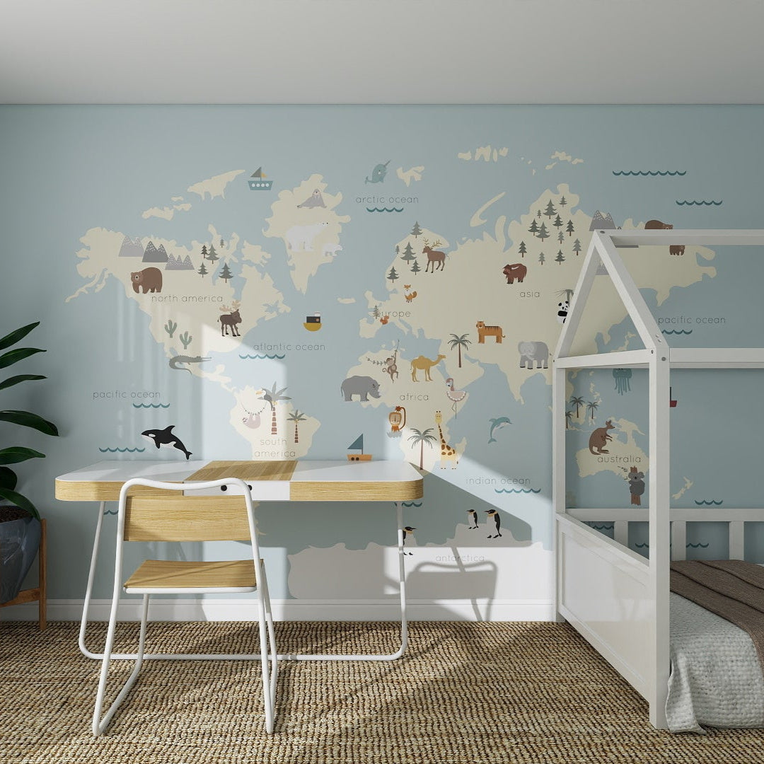 A children’s bedroom with light blue walls, a Character World Map Wallpaper Mural | Blue featuring animal illustrations, a wooden desk and chair, potted plant, white bed with brown bedding—sunlight streams in for your little adventurer.