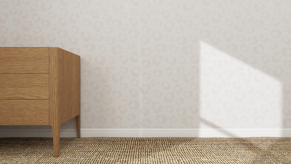 A wooden dresser with three drawers stands on a woven beige rug beside Hallie Leopard Print Wallpaper in light pink. Sunlight casts bright geometric shadows on the wall and floor.