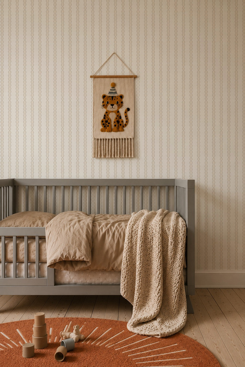 A cozy kids room features a gray crib, tan bedding, a knit blanket, a burnt orange rug, stacking toys, and charming Little Abacus Stripe Wallpaper in Off White and Taupe above the crib for a sweet finishing touch.
