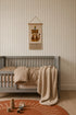 A cozy kids room features a gray crib, tan bedding, a knit blanket, a burnt orange rug, stacking toys, and charming Little Abacus Stripe Wallpaper in Off White and Taupe above the crib for a sweet finishing touch.