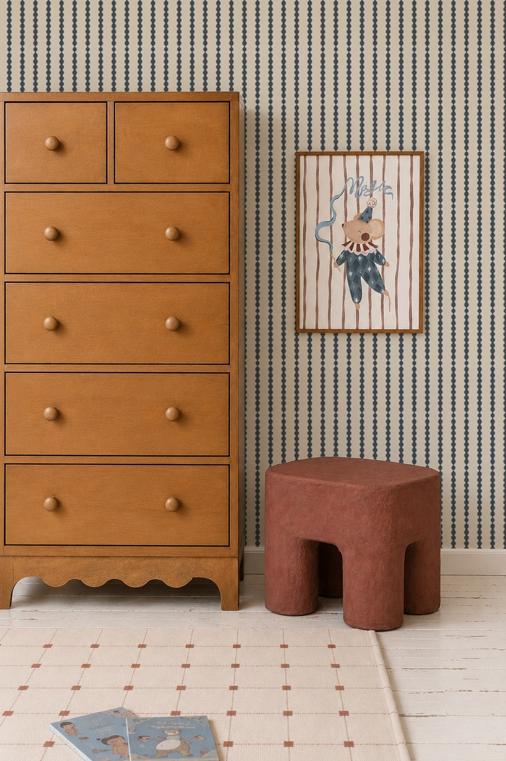 A wooden dresser stands next to a red-brown stool, with a framed animal illustration on the wall above featuring Little Abacus Stripe Wallpaper in Denim. Timeless nursery decor completes the scene, including a white grid-pattern rug and children&