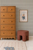 A wooden dresser stands next to a red-brown stool, with a framed animal illustration on the wall above featuring Little Abacus Stripe Wallpaper in Denim. Timeless nursery decor completes the scene, including a white grid-pattern rug and children&