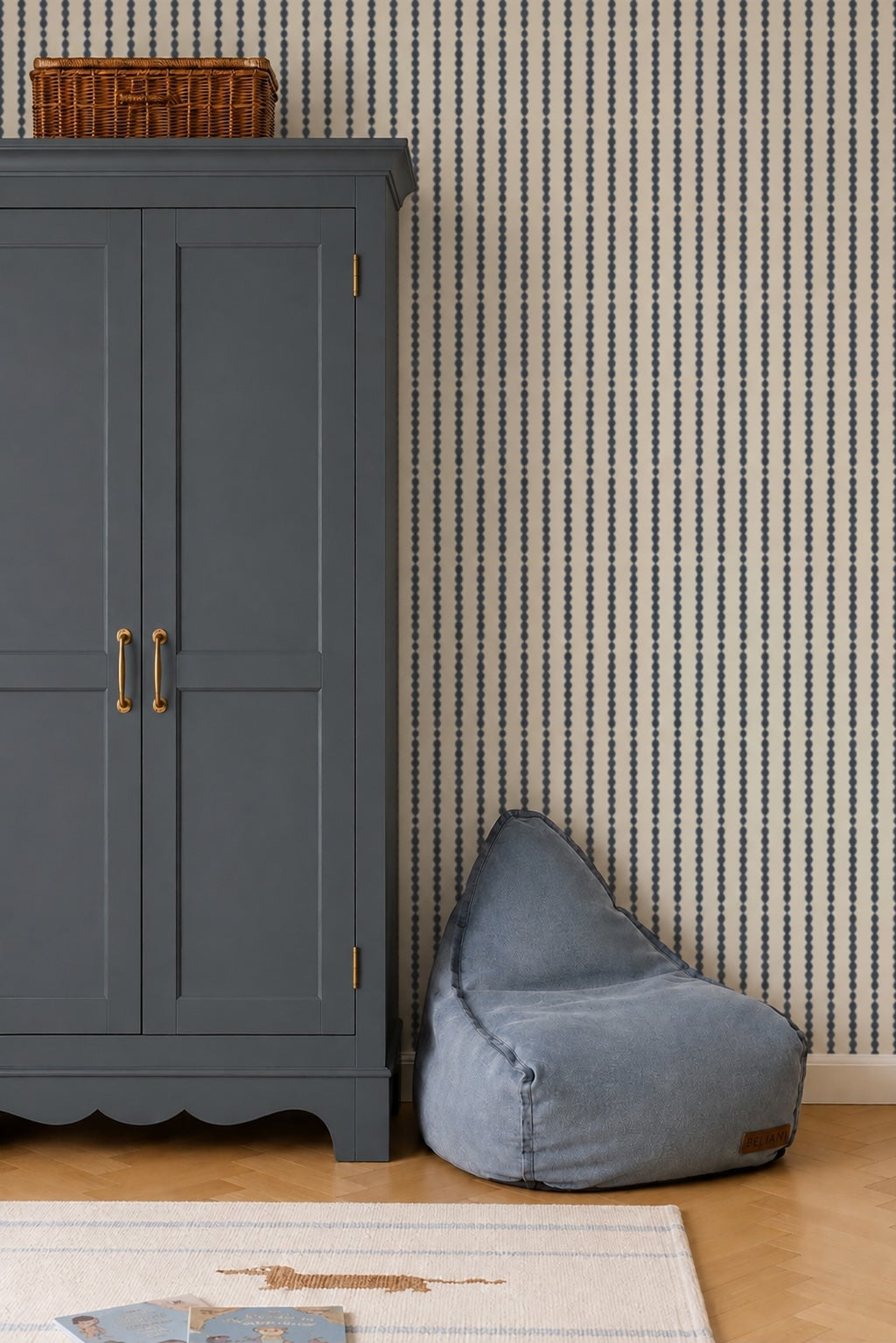 A blue wardrobe with gold handles stands beside a blue bean bag chair in a nursery featuring Little Abacus Stripe Wallpaper in Denim. A wicker basket rests atop the wardrobe, while part of a rug is visible on the wooden floor.