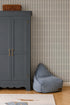 A blue wardrobe with gold handles stands beside a blue bean bag chair in a nursery featuring Little Abacus Stripe Wallpaper in Denim. A wicker basket rests atop the wardrobe, while part of a rug is visible on the wooden floor.