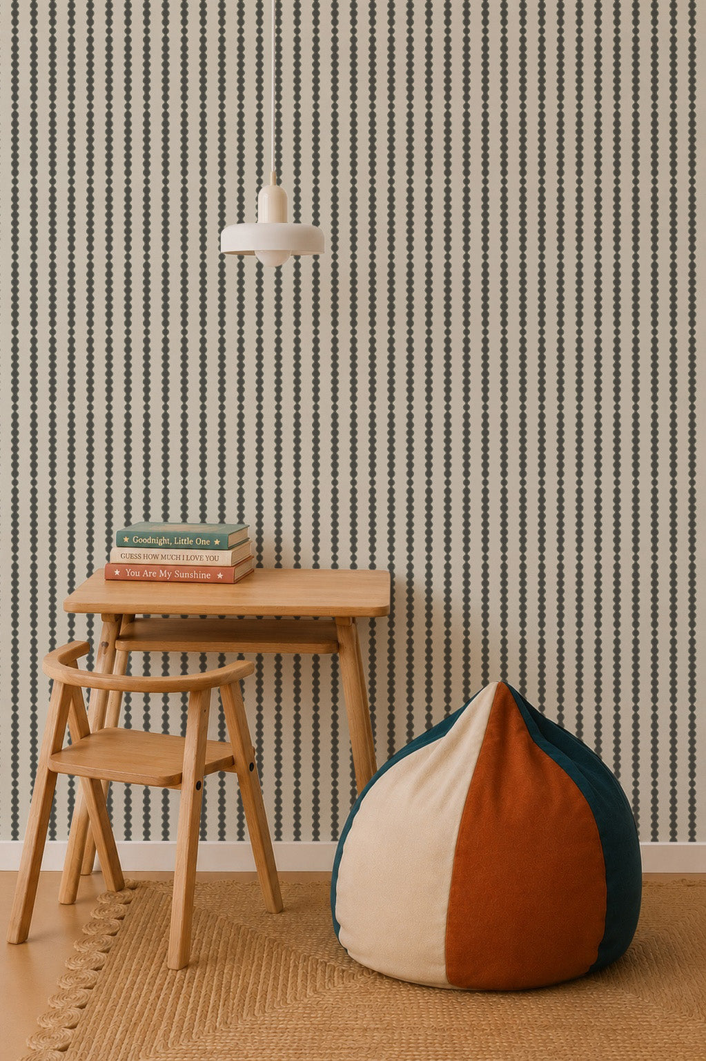 A minimalist room features a small wooden desk and chair, a stack of books, a round color-blocked bean bag, a woven rug, a pendant lamp, nursery decor for a calming vibe, and Little Abacus Stripe Wallpaper in Forest with vertical stripes on the wall.