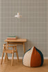 A minimalist room features a small wooden desk and chair, a stack of books, a round color-blocked bean bag, a woven rug, a pendant lamp, nursery decor for a calming vibe, and Little Abacus Stripe Wallpaper in Forest with vertical stripes on the wall.