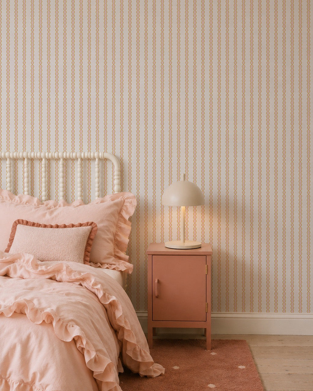 A cozy bedroom with a white spindle bed, pink ruffled bedding, pink nightstand, and beige lamp features the Little Abacus Stripe Wallpaper, Pink and Yellow—perfect nursery wallpaper for fans of soft pastel colors.