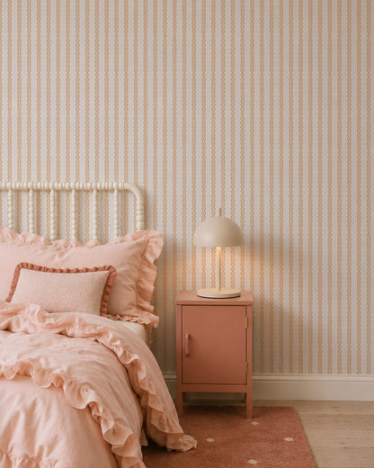 A cozy bedroom with a white spindle bed, pink ruffled bedding, pink nightstand, and beige lamp features the Little Abacus Stripe Wallpaper, Pink and Yellow—perfect nursery wallpaper for fans of soft pastel colors.