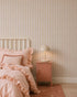 A cozy bedroom with a white spindle bed, pink ruffled bedding, pink nightstand, and beige lamp features the Little Abacus Stripe Wallpaper, Pink and Yellow—perfect nursery wallpaper for fans of soft pastel colors.