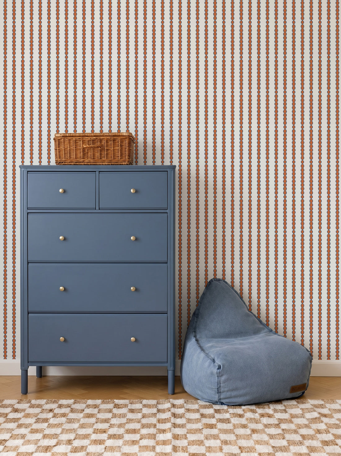 A blue dresser with silver knobs stands beside a blue bean bag chair, topped with a small wicker basket. The room features Little Abacus Stripe Wallpaper in red and blue and a checkered rug on wooden floors.