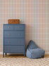 A blue dresser with silver knobs stands beside a blue bean bag chair, topped with a small wicker basket. The room features Little Abacus Stripe Wallpaper in red and blue and a checkered rug on wooden floors.