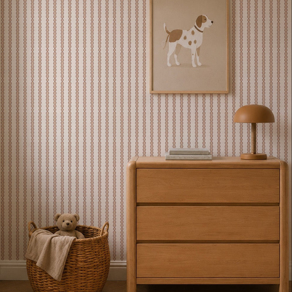 A cozy nursery with a wooden dresser, brown lamp, dog illustration, and a woven basket with a teddy bear and beige blanket, featuring Little Abacus Stripe Wallpaper in Rose.